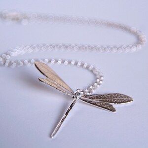 silver dragonfly necklace with leaf textured wings lies on a white surface, viewed from the insect's long body, upwards.