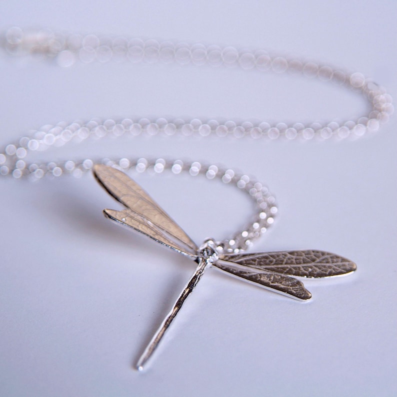 silver dragonfly necklace with leaf textured wings lies on a white surface, viewed from the insect's long body, upwards.