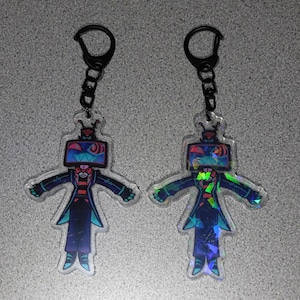 May include: Two clear acrylic keychains featuring a cartoon character with a TV head and a black chain. The character has a red and black outfit with blue and green accents. The TV screen displays a colorful design.