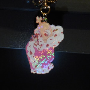 May include: A pink and white charm featuring a cartoon character wearing a crown and a pink dress. The charm has a gold-coloured keychain attachment. The character is surrounded by white fluffy clouds and hearts.
