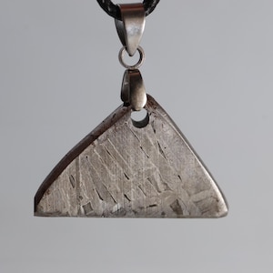 Puede incluir: Un colgante triangular hecho de un material metálico texturizado, suspendido de un cordón negro. El colgante tiene una fianza plateada y un agujero cerca de la parte superior. La superficie del colgante tiene un patrón de líneas y marcas.