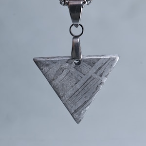 Puede incluir: Un colgante triangular de metal gris texturizado, suspendido de una cadena plateada. El colgante tiene un patrón único de líneas entrecruzadas y está unido a una fianza y cadena plateadas.