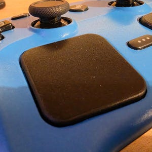 Könnte beinhalten: Ein hellblauer Handheld-Gaming-Controller mit einem großen schwarzen quadratischen Trackpad, einem schwarzen Analogstick mit strukturierter Oberfläche, einem schwarzen Steuerkreuz und einer kleinen schwarzen Taste mit drei weißen Punkten.