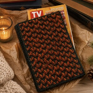 May include: A rectangular book cover featuring a textured crochet pattern in alternating dark green and burnt orange yarn. The cover is placed over a TV Guide magazine, with a lit candle in a glass holder and books visible in the background.