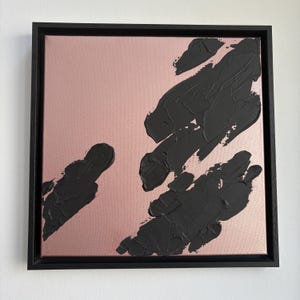 May include: Abstract art piece featuring a square canvas with a rose gold background. Bold, textured black brushstrokes are applied across the surface. The artwork is framed in a simple black wooden frame.