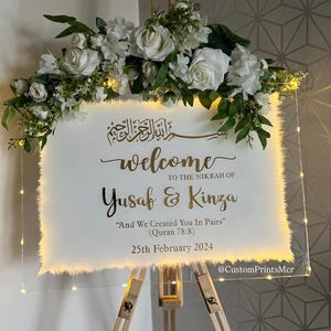 May include: A rectangular clear acrylic welcome sign for a Nikkah ceremony, featuring gold text, white floral arrangements, and warm LED fairy lights. The text reads: Welcome to the Nikkah of Yusaf & Kinza, And We Created You In Pairs, 25th February 2024.