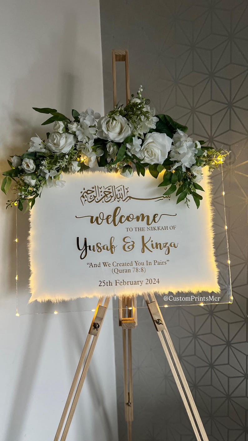 May include: A rectangular clear acrylic welcome sign for a Nikkah ceremony, featuring gold text, white floral arrangements, and warm LED fairy lights. The text reads: Welcome to the Nikkah of Yusaf & Kinza, And We Created You In Pairs, 25th February 2024.