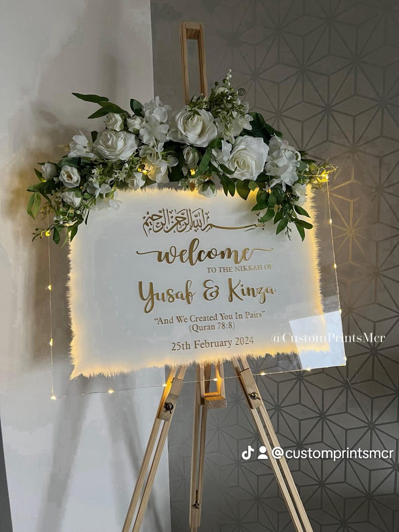 May include: A rectangular clear acrylic wedding welcome sign on a wooden easel, featuring gold calligraphy text that reads: Welcome to the Nikkah of Yusuf & Kinza, And We Created You In Pairs (Quran 78:8), 25th February 2024. Topped with white roses.