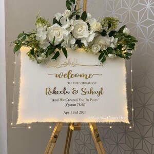 May include: A rectangular clear acrylic welcome sign on a wooden easel, adorned with white roses and greenery. The sign features gold text: "welcome TO THE NIKKAH OF Raheela & Suhayl "And We Created You In Pairs" (Quran 78:8) April 3rd 2026".