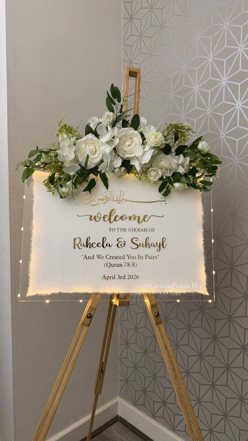 May include: A rectangular clear acrylic welcome sign on a wooden easel, adorned with white roses and greenery. The sign features gold text: "welcome TO THE NIKKAH OF Raheela & Suhayl "And We Created You In Pairs" (Quran 78:8) April 3rd 2026".