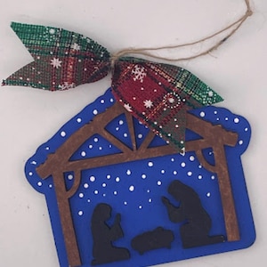 May include: A blue wooden Christmas ornament shaped like a stable, with a brown wooden frame and silhouettes of Mary, Joseph, and baby Jesus. The ornament is decorated with white dots and a plaid ribbon bow.