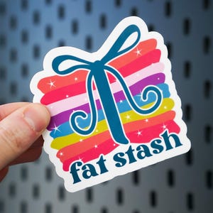 May include: A colorful sticker with the text "fat stash". The sticker has a rainbow background with a teal bow and swirls. The sticker is held by a hand.