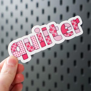May include: A sticker with the word "quilter" in pink, patchwork-style letters. The sticker is held against a gray background with a pattern of small, evenly spaced holes.