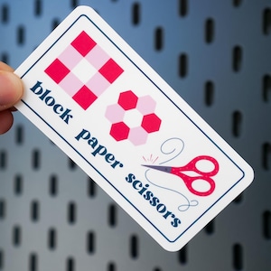 May include: A rectangular sticker with a white border features the text "block paper scissors" in navy blue. The sticker includes red and pink geometric shapes and a pair of red scissors with a navy blue handle.