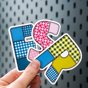 May include: A hand holds three colorful, patchwork-style stickers spelling out "& S P". The stickers feature blue, pink, and yellow sections with gingham and plaid patterns. The background is a gray surface with a pattern of small, dark rectangles.