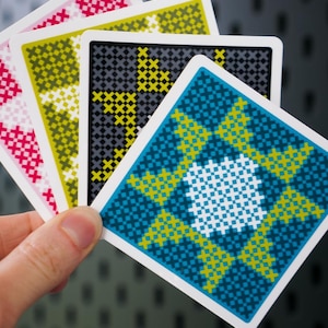 May include: A hand holding four square cards with cross-stitch patterns. The cards feature designs in pink, yellow, black, and blue, with white borders. The background is a textured gray surface.