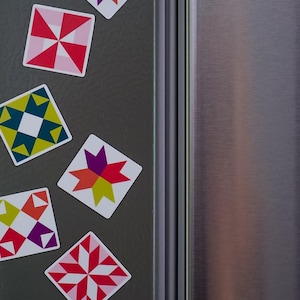 May include: A collection of square magnets featuring colorful geometric quilt block patterns in shades of red, pink, yellow, green, blue, and purple. Each magnet displays a distinct star or pinwheel design arranged on a dark gray surface.