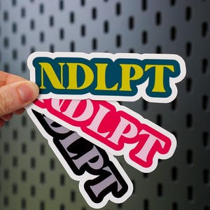 May include: Three stickers with the text "NDLPT" and "DLPT". The top sticker has yellow text with a blue outline, the middle sticker has pink text with a white outline, and the bottom sticker has gray text with a black outline.