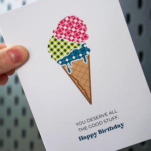 May include: A white greeting card featuring a graphic of an ice cream cone with three scoops in pink, lime green, and blue, each with a checkered pattern. The text reads: YOU DESERVE ALL THE GOOD STUFF. Happy Birthday in black and blue lettering.