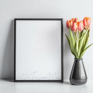 May include: A black picture frame with a white background sits on a white surface next to a vase of orange tulips.