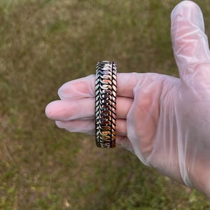 May include: A silver and gold-toned bracelet with a woven design. The bracelet is held in a gloved hand against a backdrop of green grass. The bracelet's intricate pattern is visible, showcasing its detailed craftsmanship.