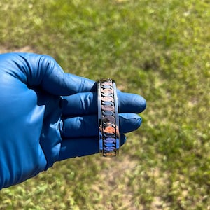 May include: A silver-toned bracelet with a repeating pattern of interlocking shapes. The bracelet reflects shades of blue, orange, and purple. The bracelet is held against a background of green grass.