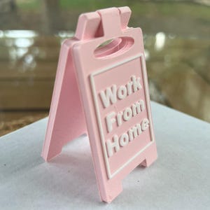 Op de afbeelding: Een lichtroze, dubbelzijdig bord met de woorden "Work From Home" in witte blokletters. Het bord is in A-frame stijl en lijkt van plastic te zijn. Het bord staat op een wit oppervlak.