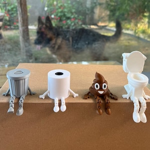 Puede incluir: Un conjunto de figuras caprichosas impresas en 3D: un cubo de basura gris, un rollo de papel higi&eacute;nico blanco, un emoji de caca marr&oacute;n sonriente y un inodoro blanco. Cada figura tiene brazos y piernas, sentada sobre una superficie marr&oacute;n.