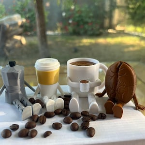 Może przedstawiać: Kapryśna aranżacja o tematyce kawowej z srebrnym ekspresem do kawy, kubkiem na wynos z żółtą osłonką, białym kubkiem do kawy i postacią ziarna kawy. Ziarna kawy są rozrzucone wokół figurek. Tło przedstawia rozmyty widok na zewnątrz.