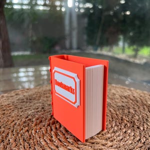 Puede incluir: Un sujetalibros naranja con forma de libro con la palabra "Bookmarks" en una etiqueta blanca. El soporte est&aacute; lleno de marcadores y se encuentra sobre una superficie tejida.