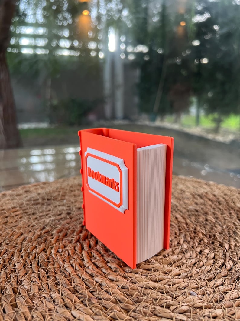 Puede incluir: Un sujetalibros naranja con forma de libro con la palabra "Bookmarks" en una etiqueta blanca. El soporte est&aacute; lleno de marcadores y se encuentra sobre una superficie tejida.
