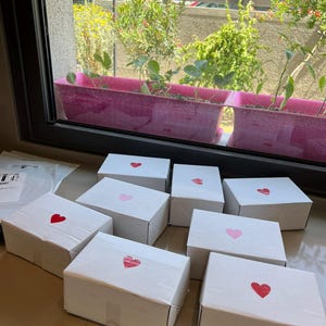 Puede incluir: Una colecci&oacute;n de cajas de cart&oacute;n blancas, cada una adornada con una pegatina en forma de coraz&oacute;n en varios tonos de rojo y rosa. Las cajas est&aacute;n dispuestas sobre una encimera, con una ventana y plantas en el fondo.