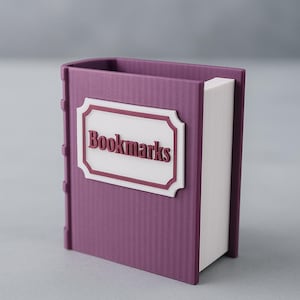 Può includere: Un portasegnapagina viola a forma di libro, con la scritta "Bookmarks" sul davanti. Il supporto ha un design strutturato e a coste e un'etichetta bianca con un bordo marrone. Le pagine sono bianche.