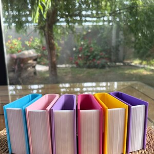 Puede incluir: Una colecci&oacute;n de cajas de almacenamiento con forma de libro de colores. Las cajas son en tonos de azul, rosa, morado y amarillo, con p&aacute;ginas blancas. Est&aacute;n dispuestas sobre una superficie tejida, con un fondo exterior borroso.