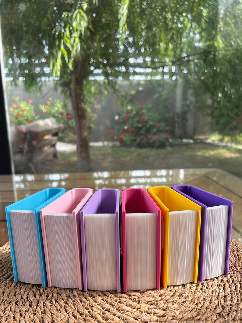 Puede incluir: Una colecci&oacute;n de cajas de almacenamiento con forma de libro de colores. Las cajas son en tonos de azul, rosa, morado y amarillo, con p&aacute;ginas blancas. Est&aacute;n dispuestas sobre una superficie tejida, con un fondo exterior borroso.