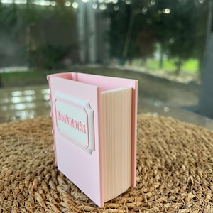 Può includere: Un organizer da scrivania a forma di libro rosa con la scritta "Bookmarks" in una cornice bianca decorativa. L'organizer ha un esterno rosa chiaro e un interno beige, che ricorda le pagine di un libro. L'articolo è posto su una superficie marrone intrecciata.