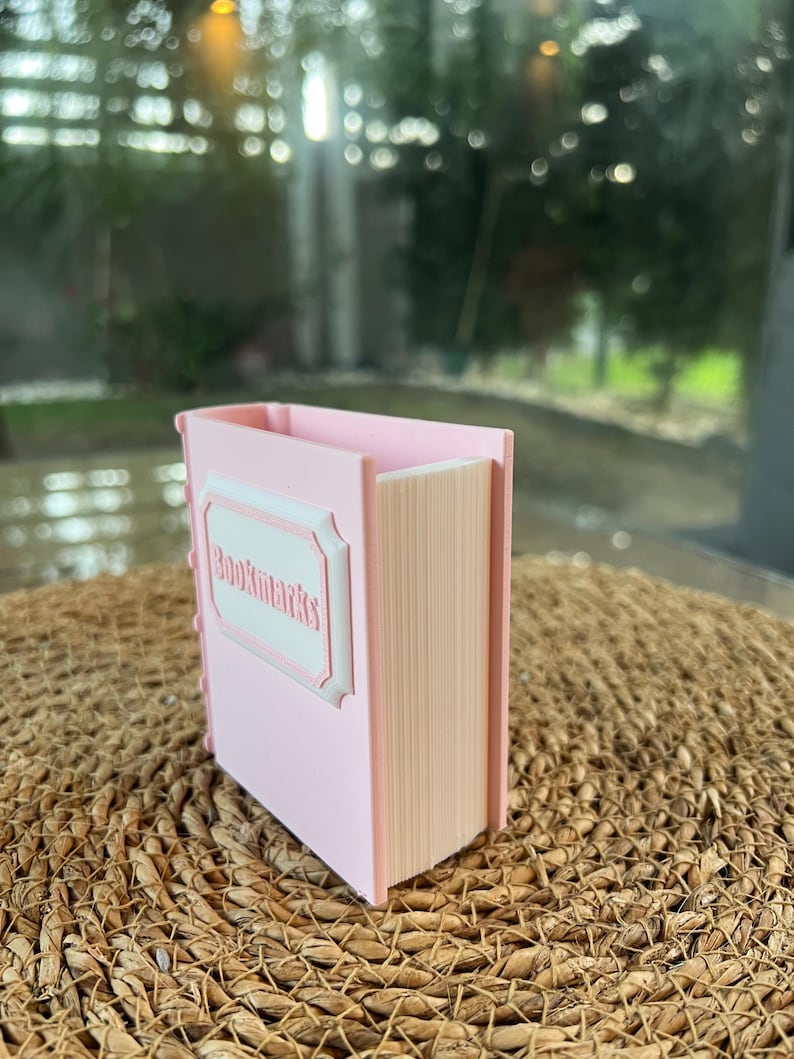 Puede incluir: Un organizador de escritorio rosa con forma de libro con la palabra "Bookmarks" en un marco blanco decorativo. El organizador tiene un exterior rosa claro y un interior beige, que se asemeja a las p&aacute;ginas de un libro. Se encuentra sobre una superficie tejida marr&oacute;n.