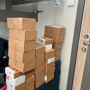 Puede incluir: Una pila de cajas de cart&oacute;n de varios tama&ntilde;os, algunas con etiquetas blancas y rojas, apiladas sobre un banco tapizado de color turquesa. Las cajas sugieren una variedad de art&iacute;culos.
