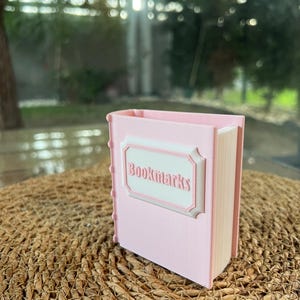 Puede incluir: Un recipiente rosa con forma de libro con la palabra "Bookmarks" en un marco blanco decorativo. El recipiente tiene un lomo con una serie de peque&ntilde;as hendiduras redondas. El fondo est&aacute; borroso, con una estera tejida natural y una ventana.