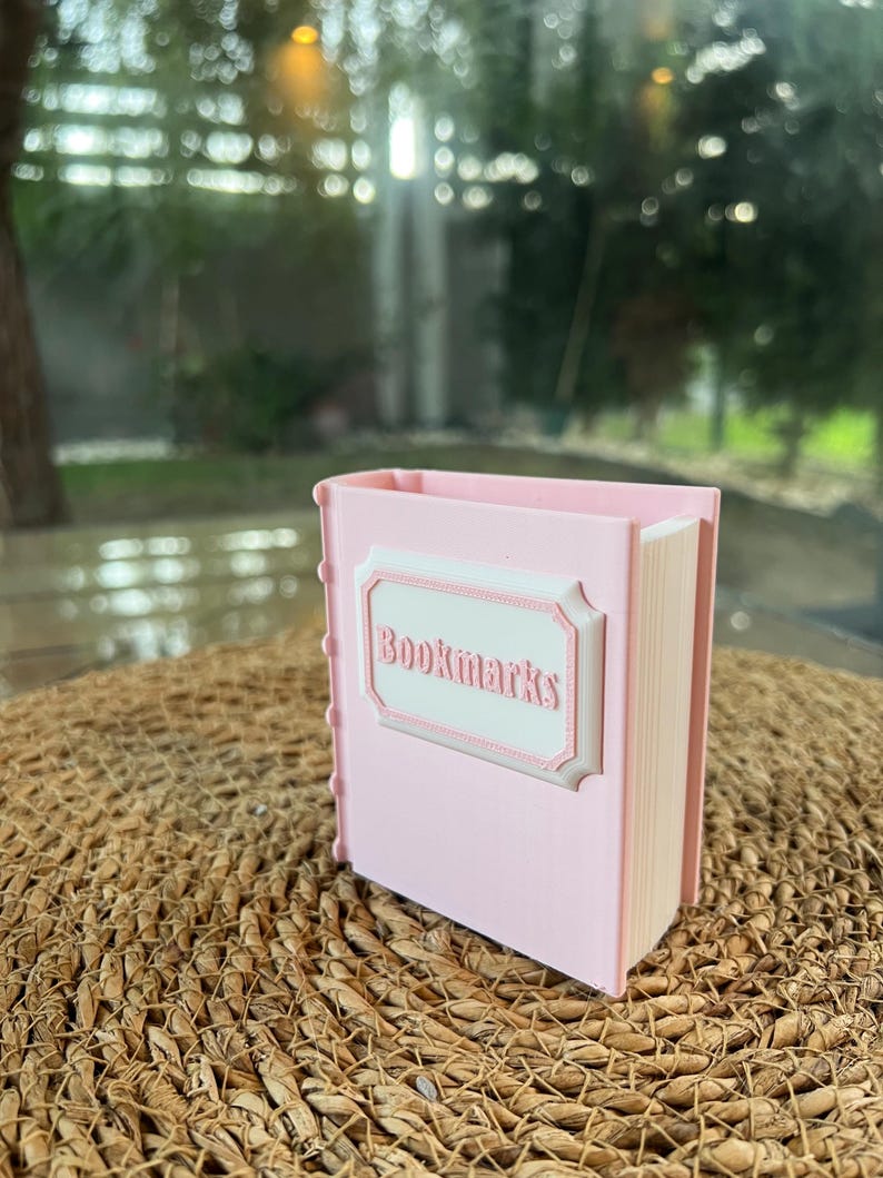 Puede incluir: Un recipiente rosa con forma de libro con la palabra "Bookmarks" en un marco blanco decorativo. El recipiente tiene un lomo con una serie de peque&ntilde;as hendiduras redondas. El fondo est&aacute; borroso, con una estera tejida natural y una ventana.