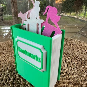 Puede incluir: Un sujetalibros verde con forma de libro con la palabra "Bookmarks" en verde. El soporte contiene marcadores con forma de gato rosa y blanco. El soporte est&aacute; sobre una superficie tejida.