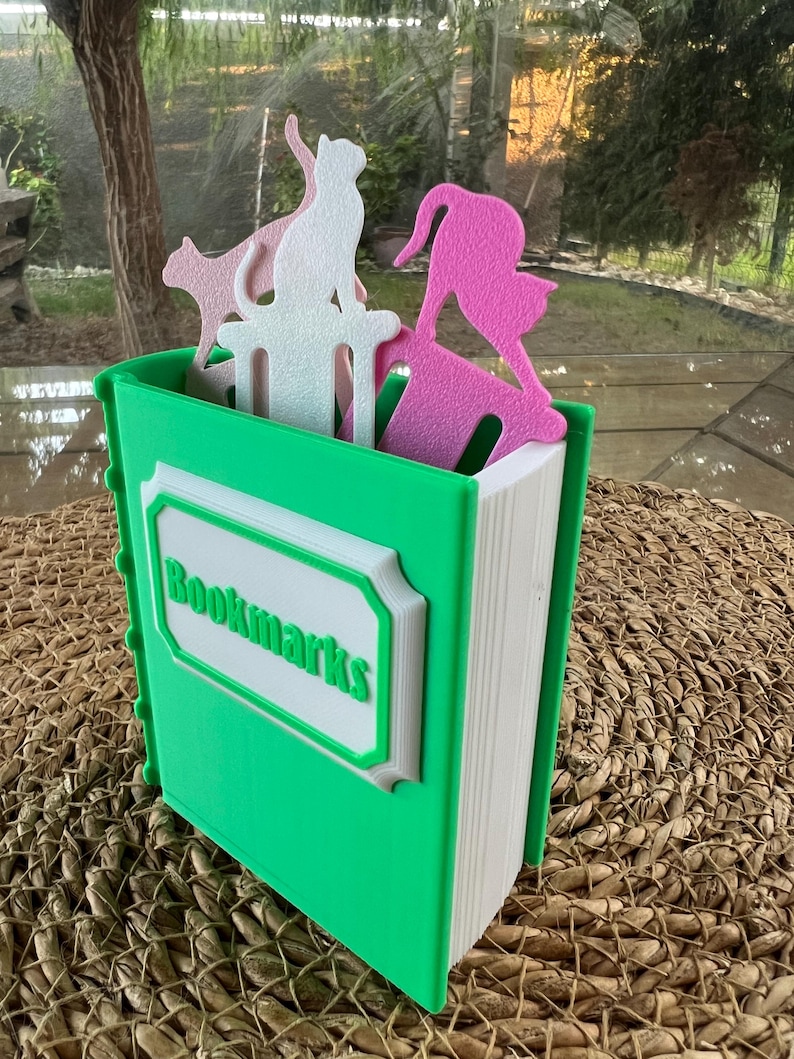 Puede incluir: Un sujetalibros verde con forma de libro con la palabra "Bookmarks" en verde. El soporte contiene marcadores con forma de gato rosa y blanco. El soporte est&aacute; sobre una superficie tejida.
