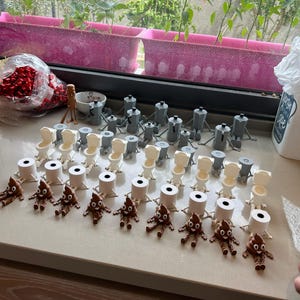 Puede incluir: Una colecci&oacute;n de figuras novedosas, que incluyen inodoros en miniatura, cafeteras y emojis de caca sonrientes. Las figuras est&aacute;n dispuestas sobre una superficie de color claro, con una ventana y plantas en el fondo.
