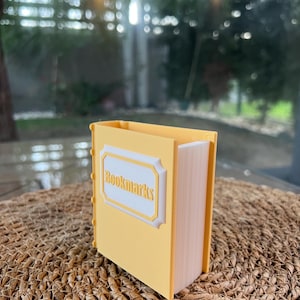 Puede incluir: Un contenedor rectangular amarillo con forma de libro, con la palabra "Bookmarks" en la parte delantera. El contenedor est&aacute; sobre una superficie tejida, con un fondo borroso de vegetaci&oacute;n.