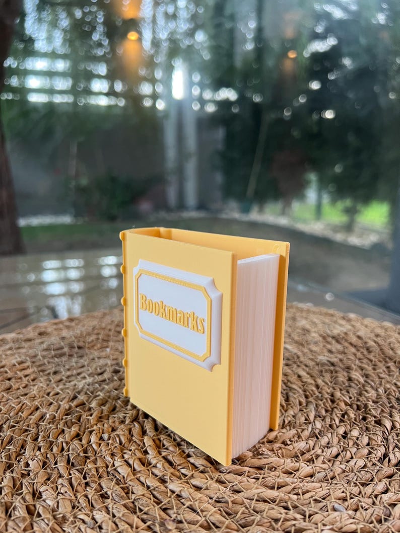 Puede incluir: Un contenedor rectangular amarillo con forma de libro, con la palabra "Bookmarks" en la parte delantera. El contenedor est&aacute; sobre una superficie tejida, con un fondo borroso de vegetaci&oacute;n.