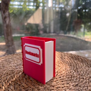 Puede incluir: Un sujetalibros rojo con forma de libro, con la palabra "Bookmarks" en un marco blanco en la portada. El sujetalibros est&aacute; lleno de p&aacute;ginas blancas y est&aacute; sobre una superficie tejida.