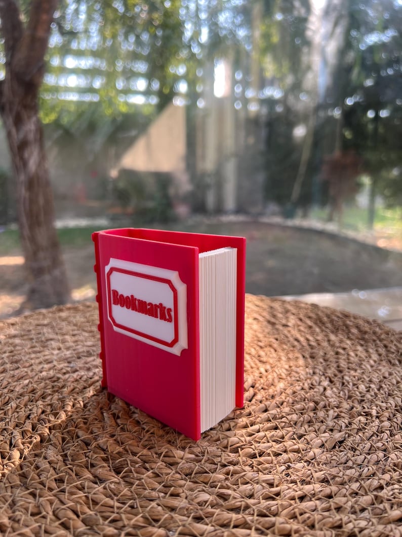 Puede incluir: Un sujetalibros rojo con forma de libro, con la palabra "Bookmarks" en un marco blanco en la portada. El sujetalibros est&aacute; lleno de p&aacute;ginas blancas y est&aacute; sobre una superficie tejida.