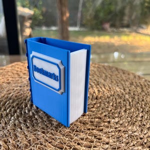 Puede incluir: Un soporte para marcap&aacute;ginas azul y blanco con forma de libro. El soporte tiene la palabra "Bookmarks" en un marco blanco en la parte delantera. El soporte est&aacute; sobre una superficie tejida.