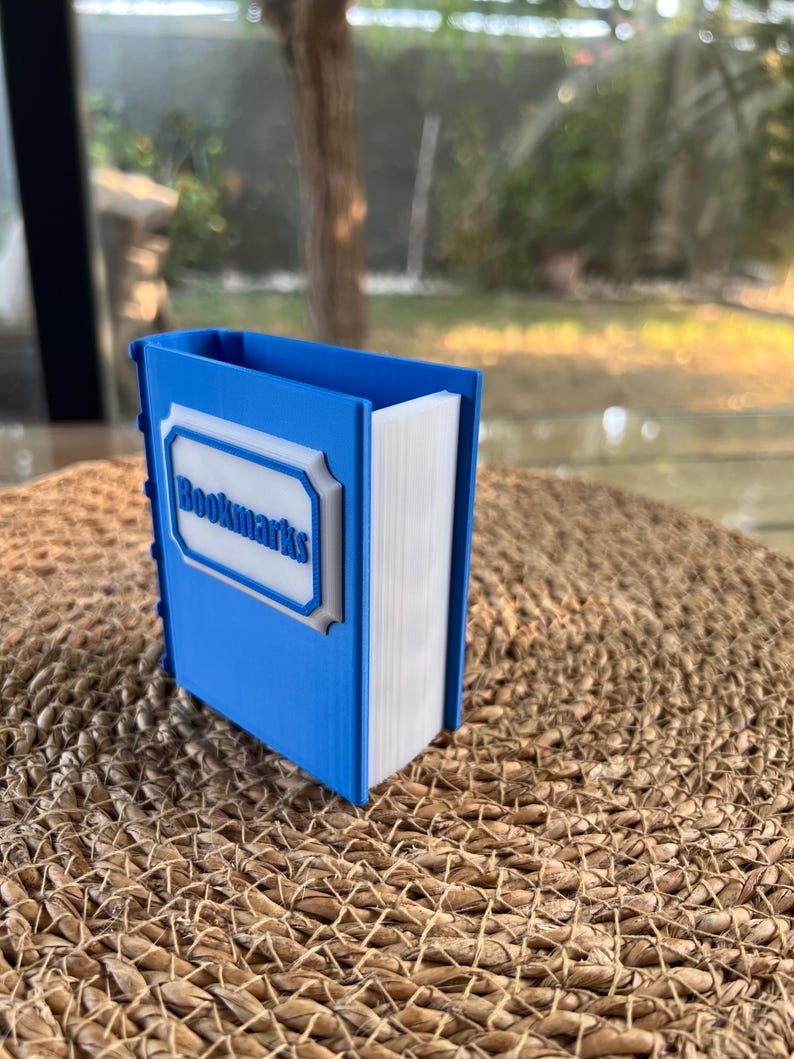 Puede incluir: Un soporte para marcap&aacute;ginas azul y blanco con forma de libro. El soporte tiene la palabra "Bookmarks" en un marco blanco en la parte delantera. El soporte est&aacute; sobre una superficie tejida.