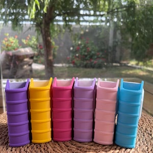 Puede incluir: Seis peque&ntilde;as macetas con forma de libro, de colores vibrantes: morado, amarillo, rosa, lavanda, rosa claro y azul. Las macetas est&aacute;n dispuestas en fila sobre una superficie tejida.